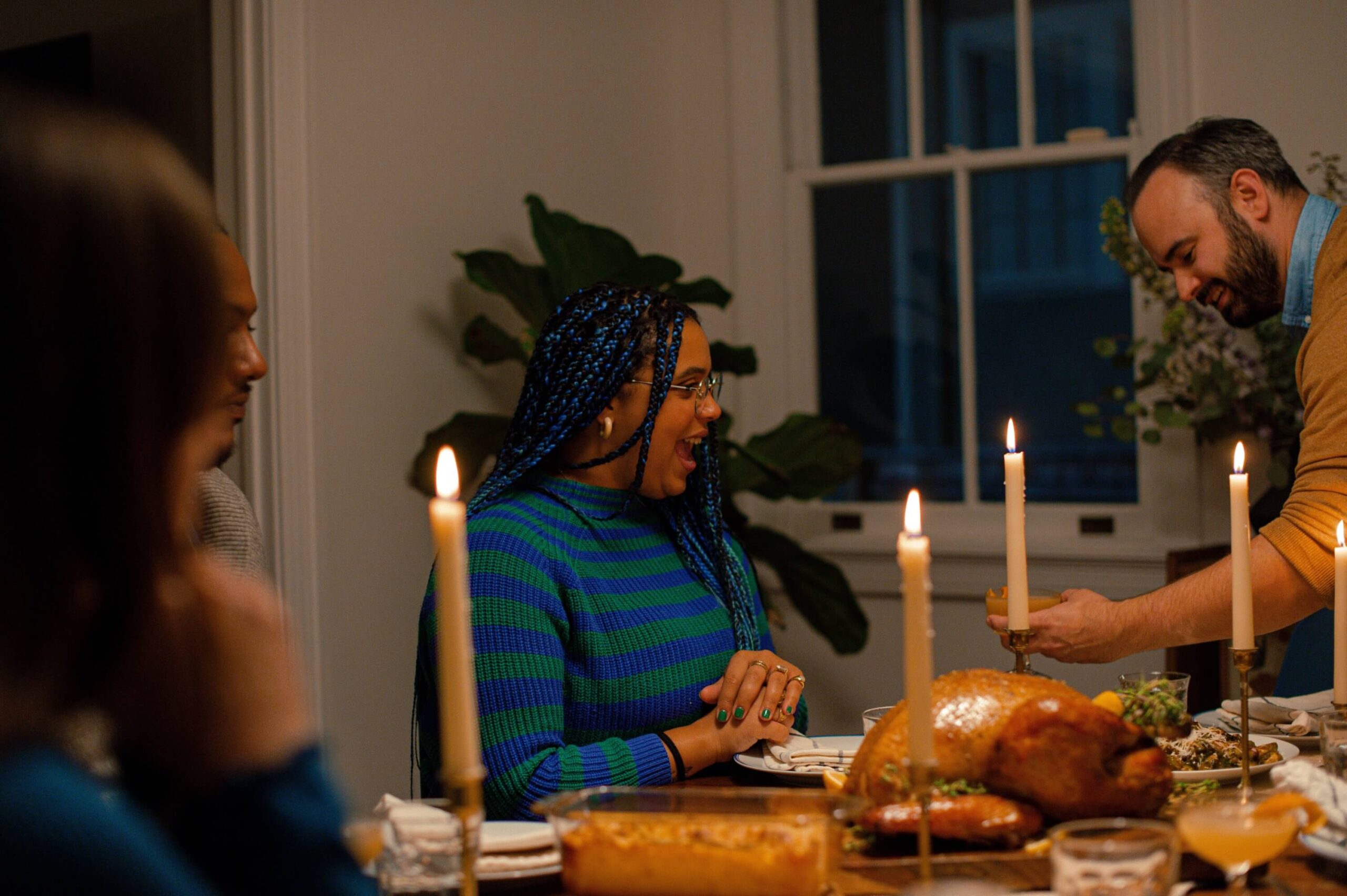 A family sits at a holiday meal surrounded by lit candles and a food spread including a turkey.
