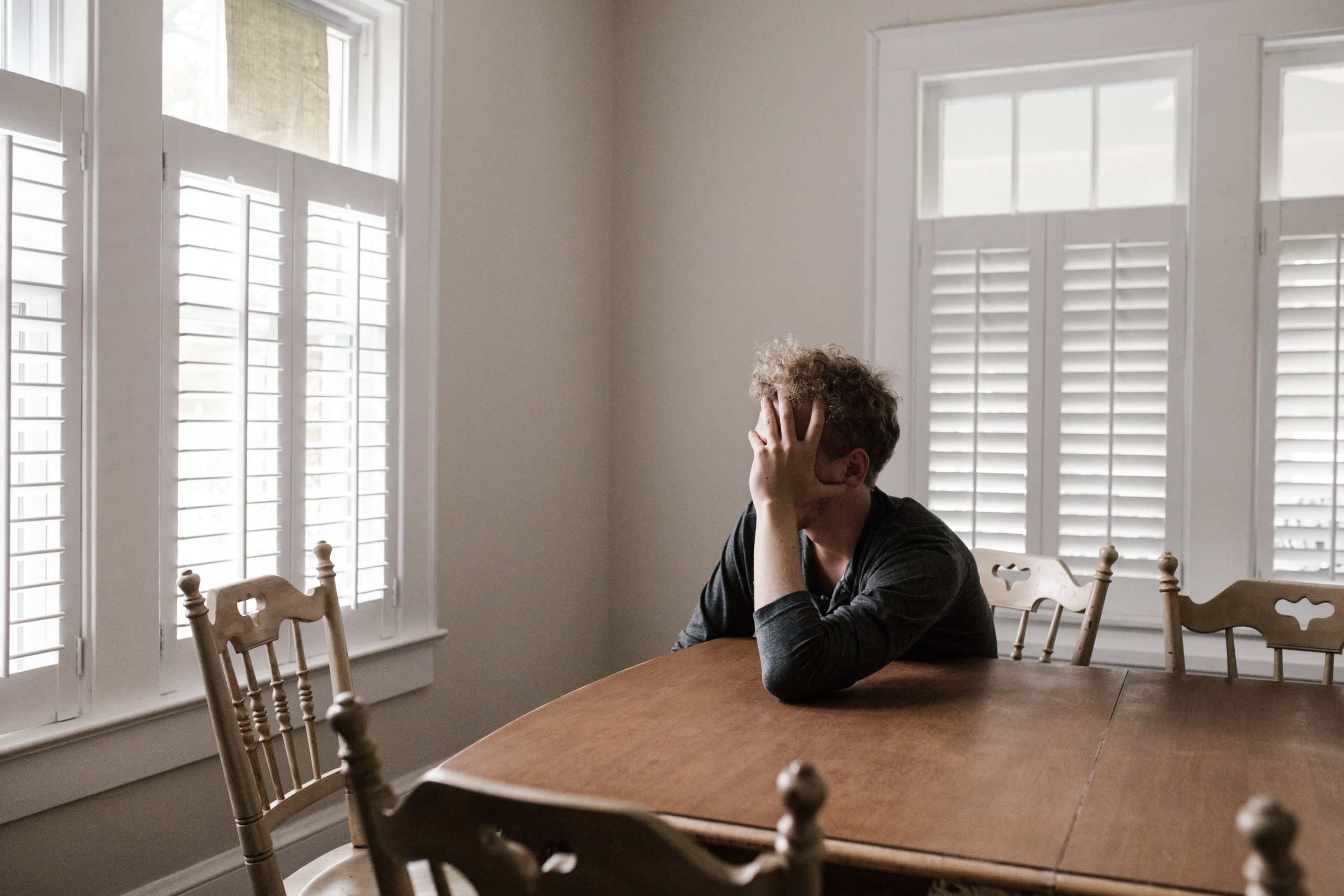 Man sitting at a table at his house with his head in his hands looking out the window