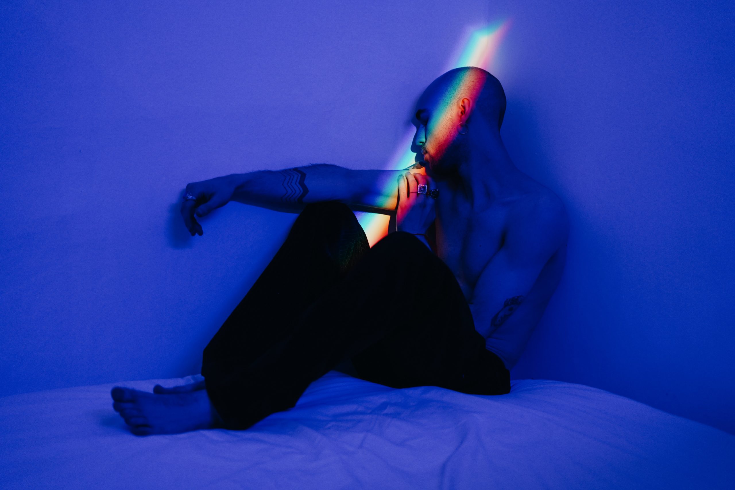 Man sitting shirtless on a bed with a rainbow light shining on him