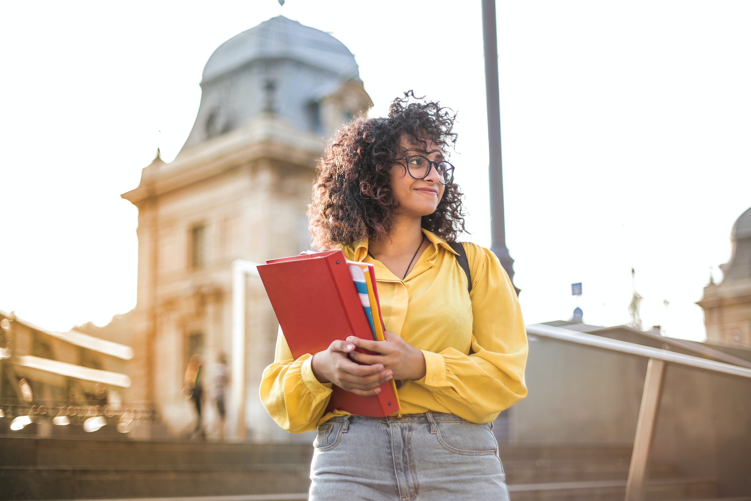 college student standing on outdoor steps, holding books and looking out with a positive facial expression