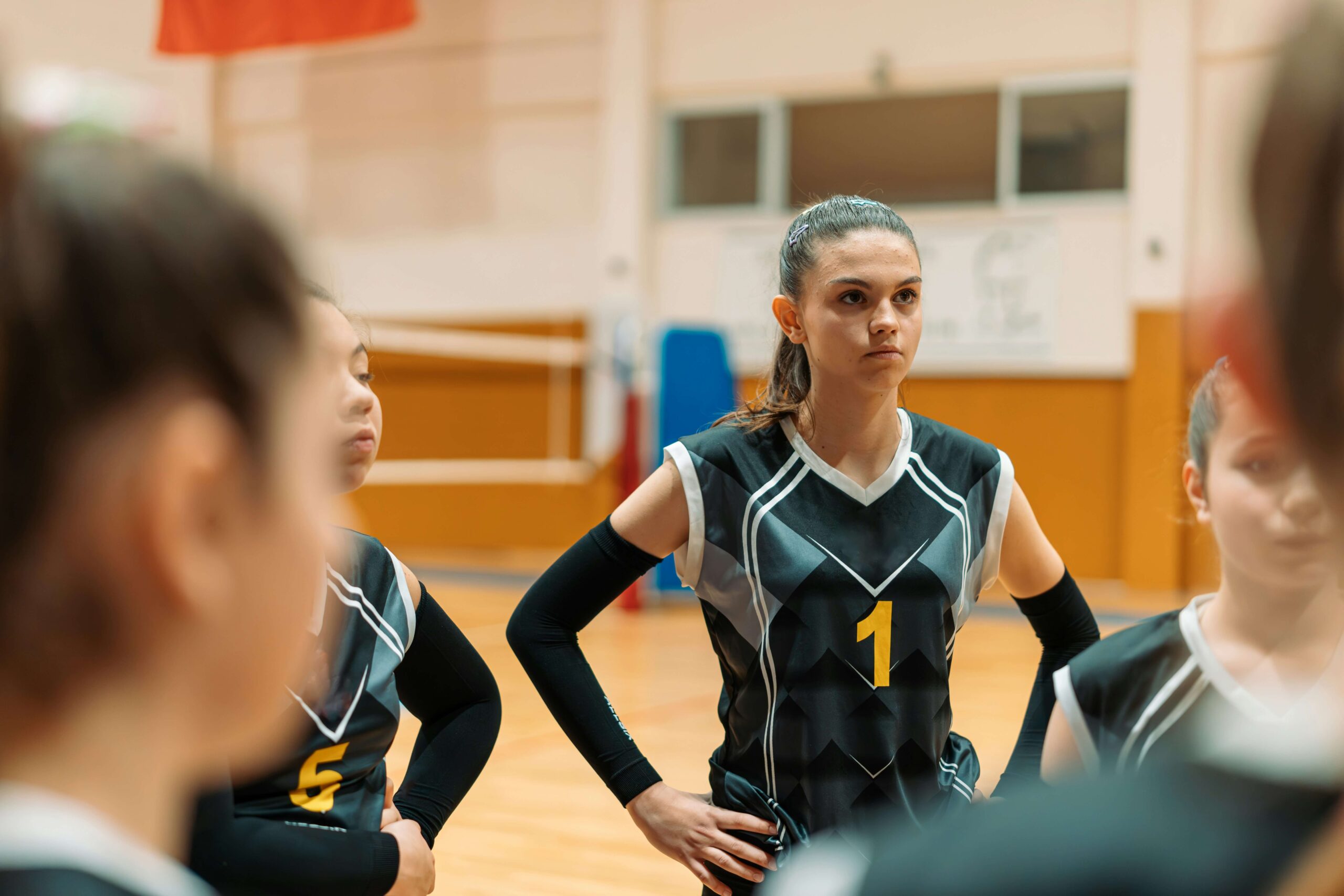 A young person standing with their hands on their hips on a volleyball court, listening to their teammates speak.