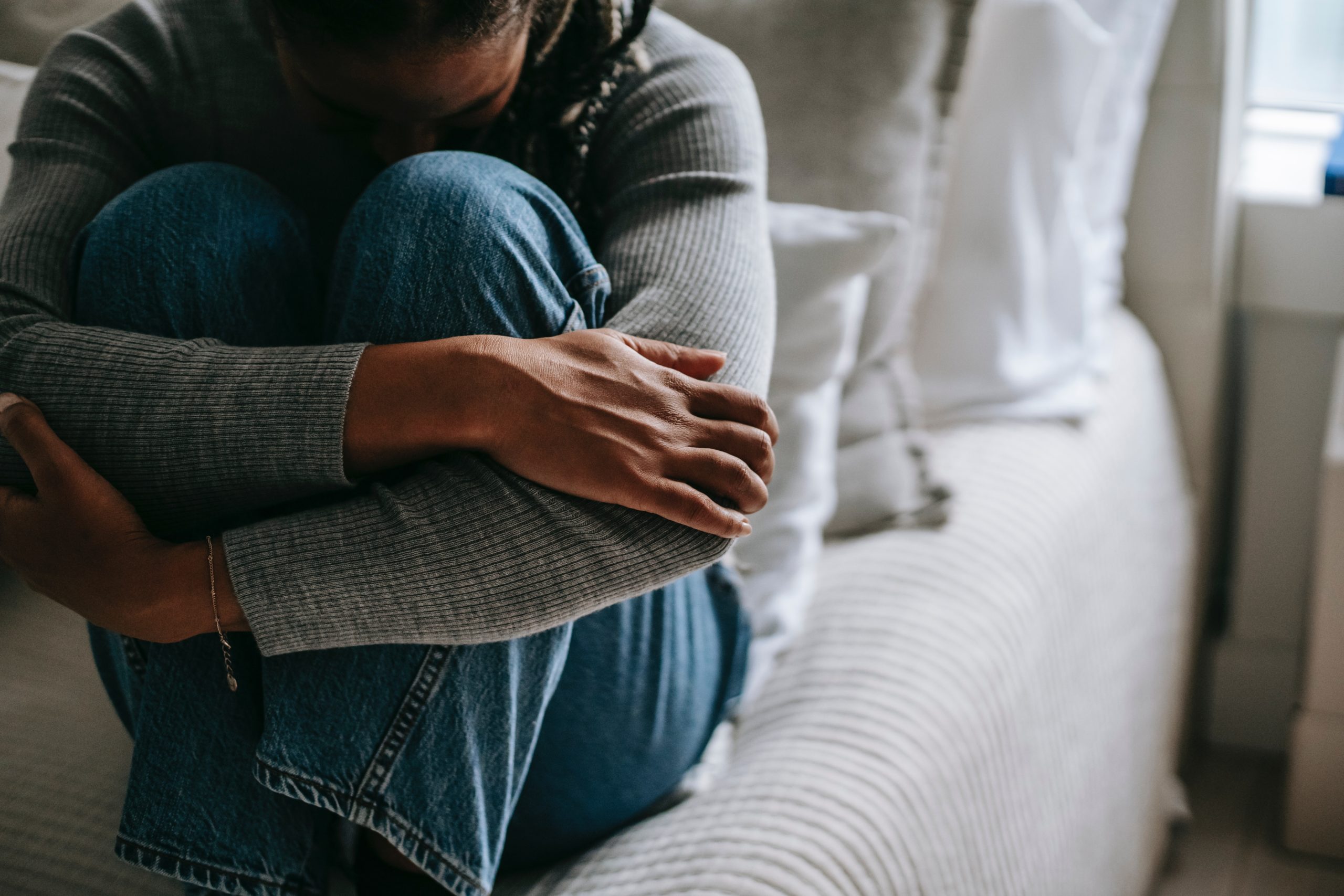 woman sitting on bed looking upset with knees up, forehead touching her knees and her arms wrapped around her legs