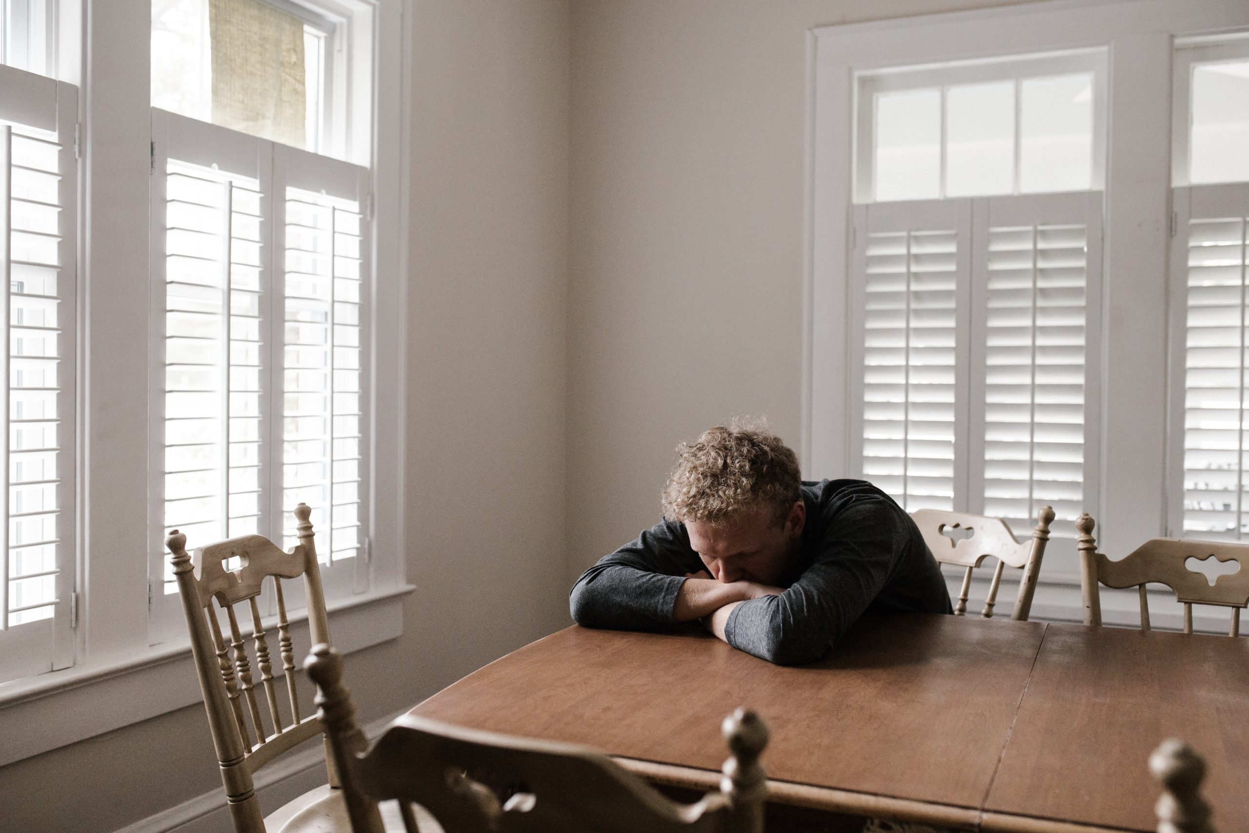 young man feeling anxious sitting at the dining room table with his face pressed into arms on the table. window blinds are open behind and to the right of him