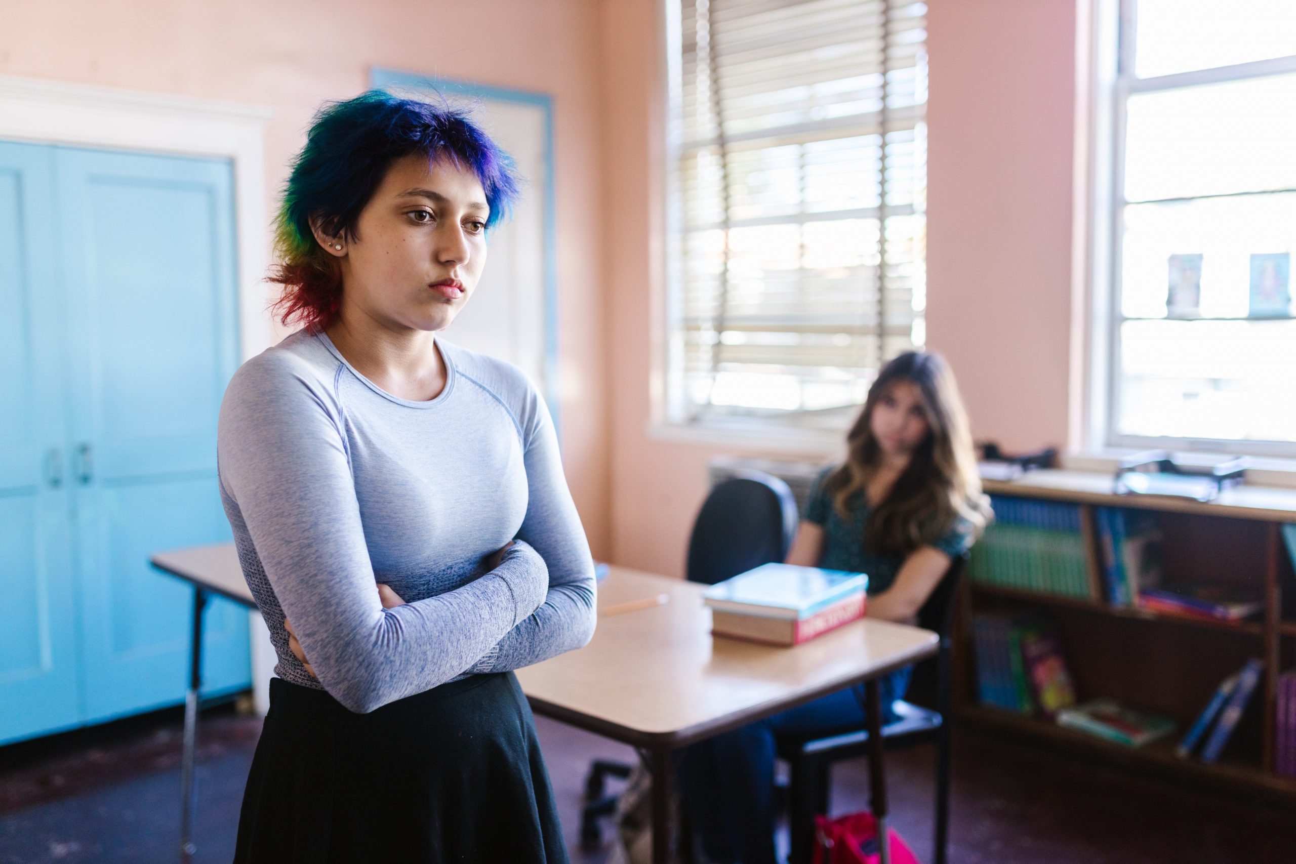 student standing in classroom with arms crossed and facial expression of sadness. second student in background with books on desk staring at student standing up. she has a somber looking facial expression.