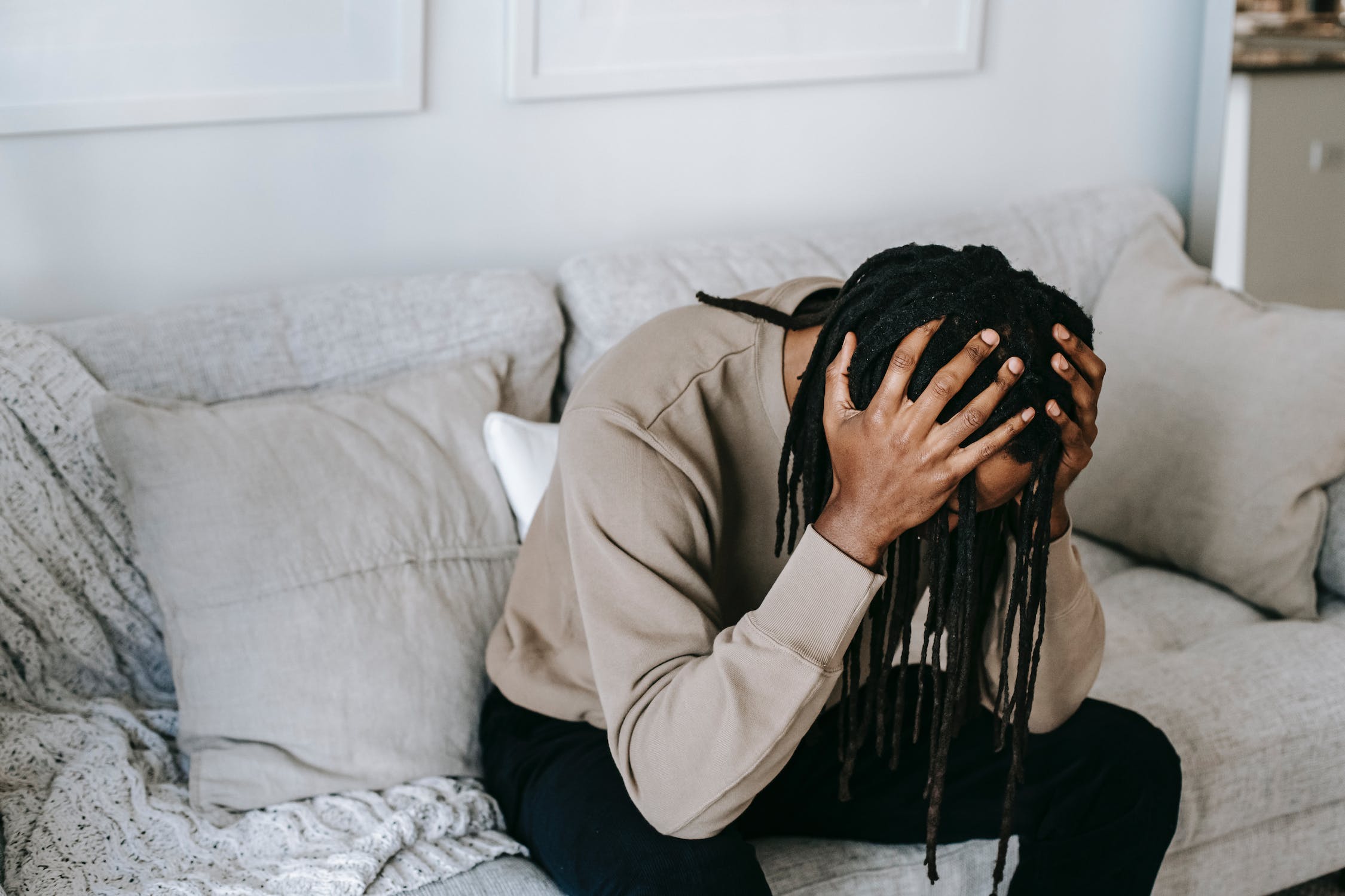 black guy with locs wearing beige sweater sitting on the light colored couch with head down and hands on his head