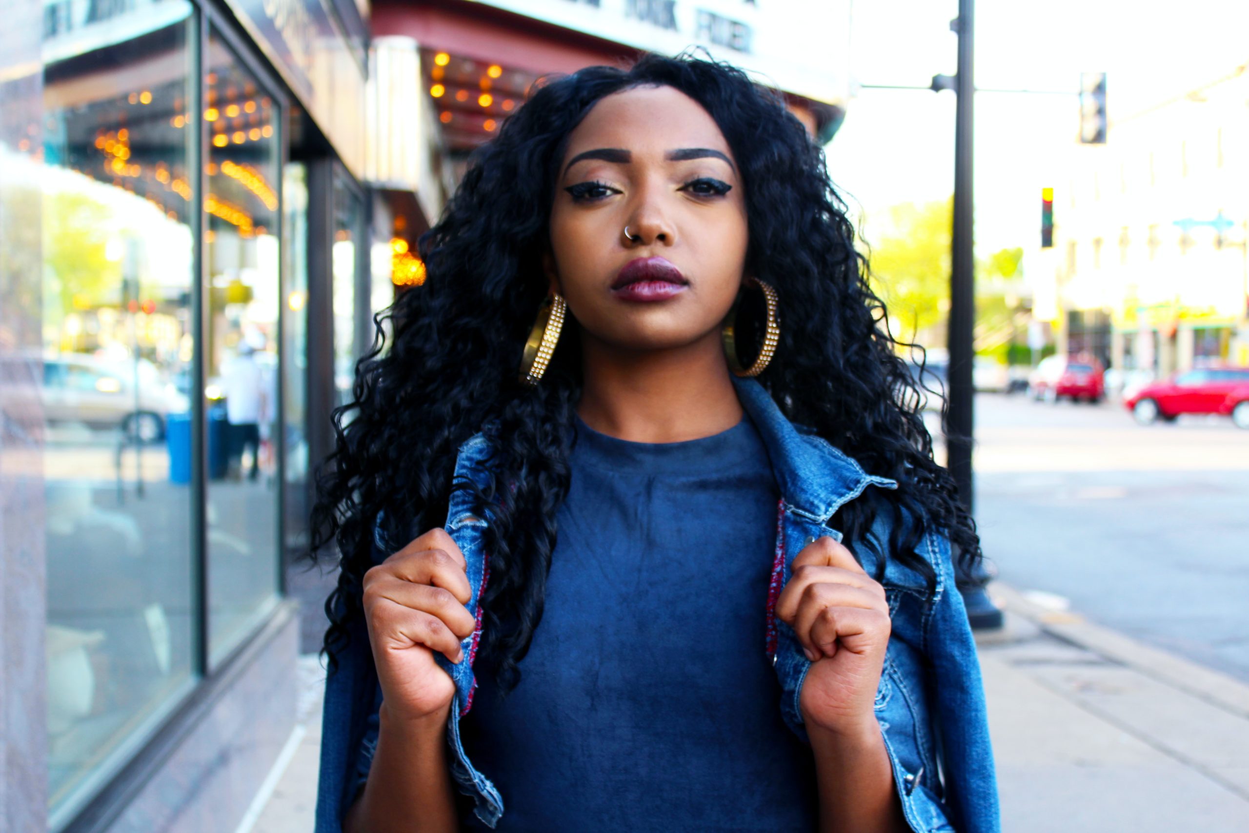 young black woman with long black curly hair wearing make-up gold earrings with both hands holding the collar of her blue denim jacket walking passed a glass window with sidewalk and street in the background