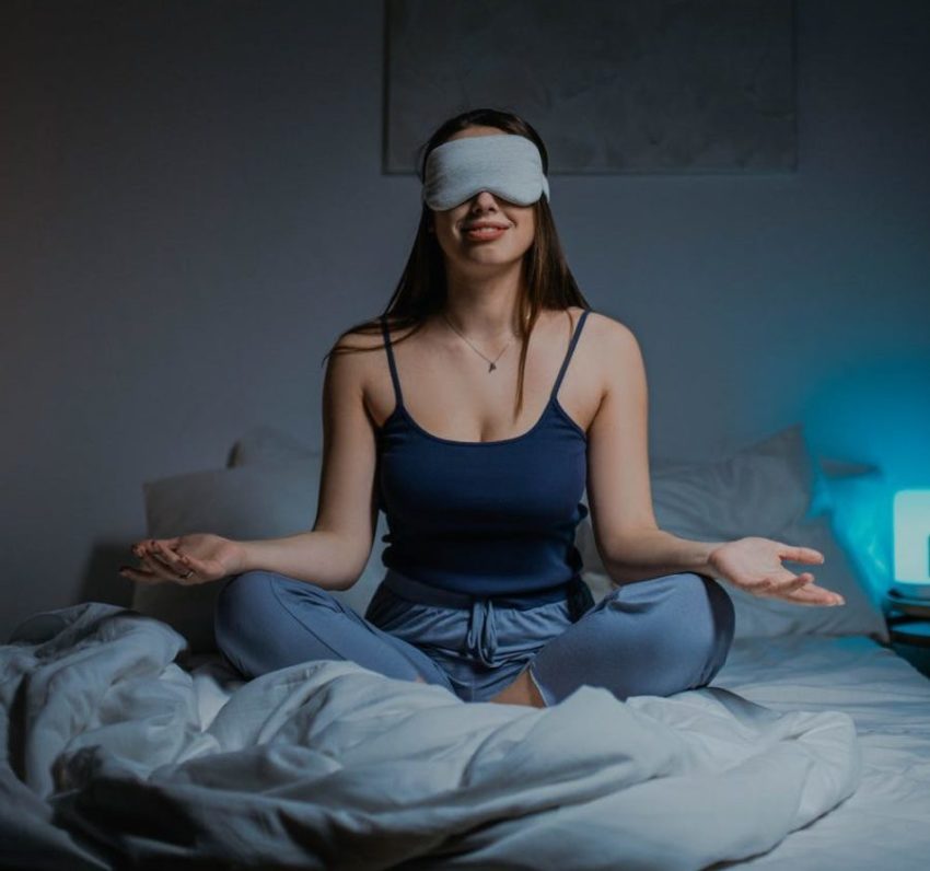 young adult with sleep mask on sitting in bed with hands on knees facing up