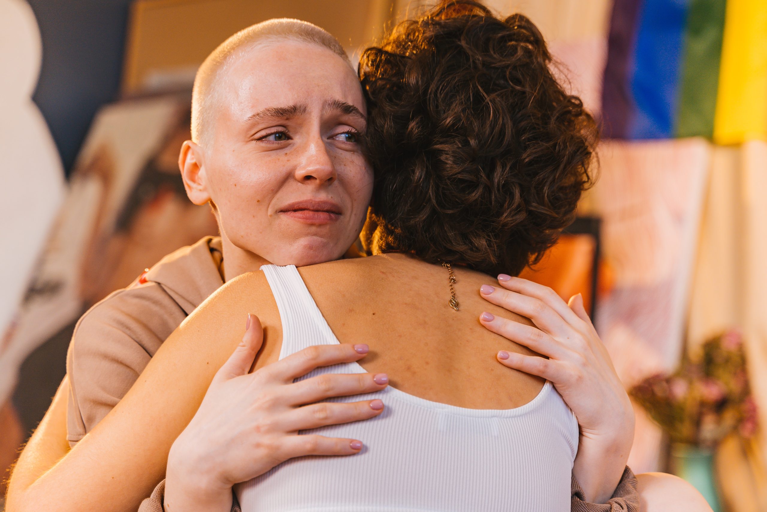 two women hugging with one woman looking upset. upset woman looks like on the verge of tears, has a buzz cut, other womens back is front facing