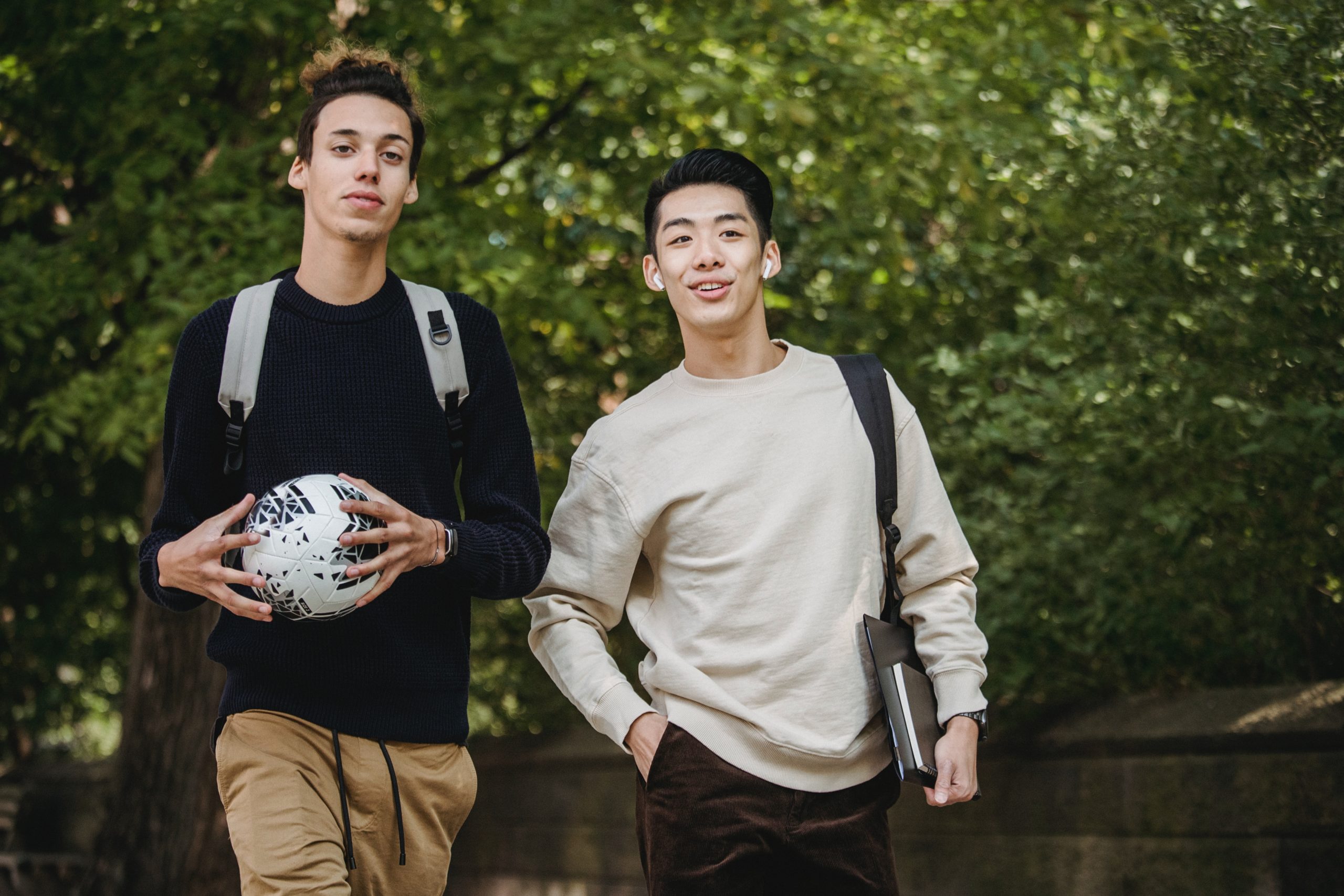 two male students walking in the park. one young man smiling with airpods in his ear and the other young man holding a soccer ball in both hands. green trees and foliage in the background