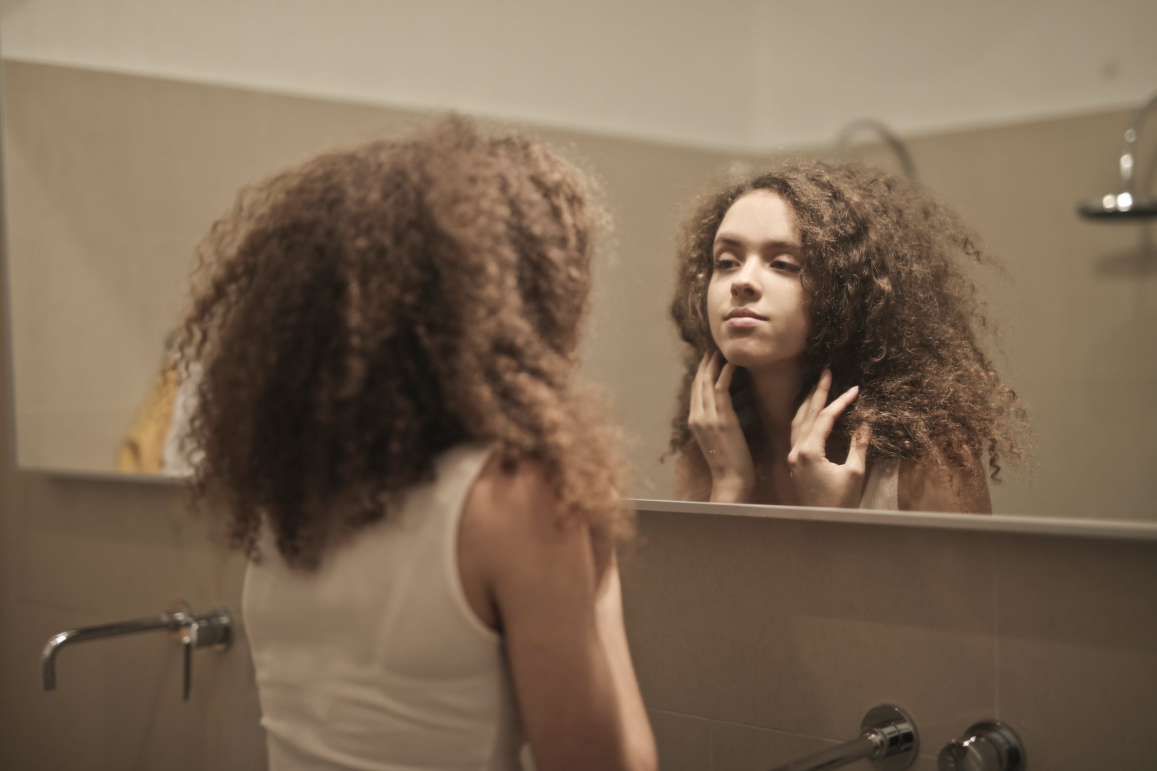 young-woman-with-thick-curly-brown-hair-looking-at-herself-in-bathroom-mirror-with-hands-by-her-face.