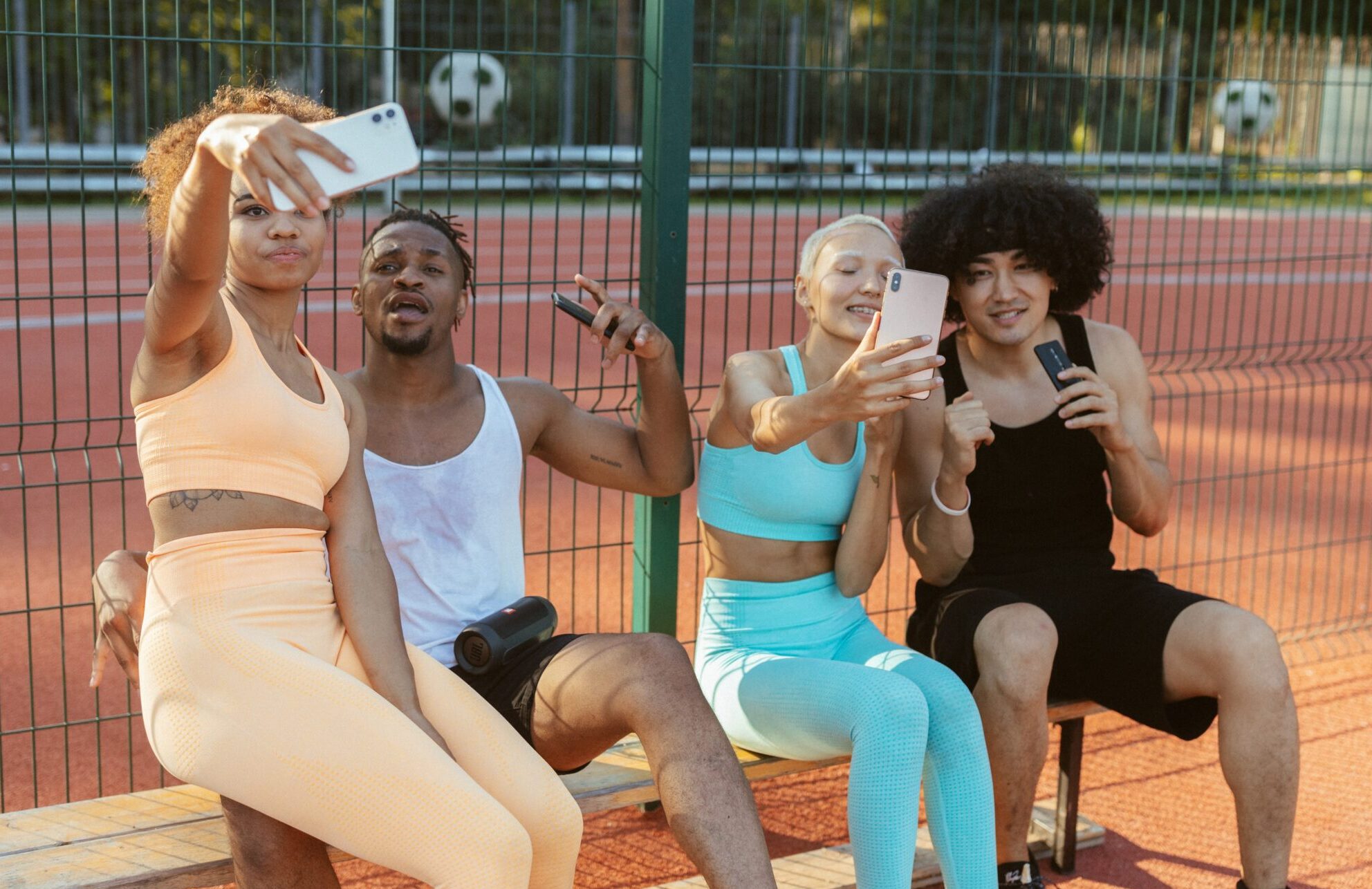 group of friends taking a selfie while sitting on an outdoor bench beside a basketball court