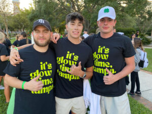 three male students hanging outside at neon nights two wearing caps and all wearing black shirts with its glow time in neon green writing