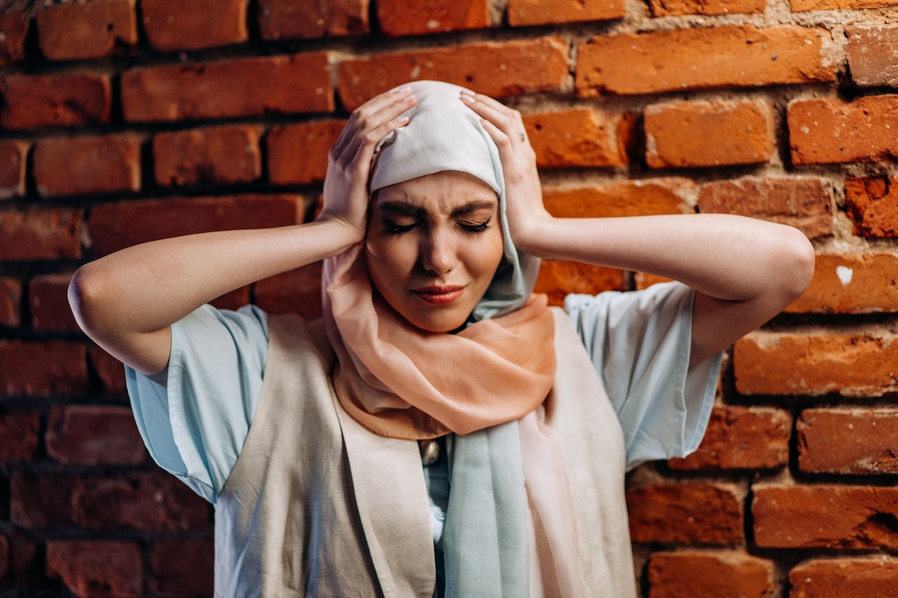 young woman of a diverse race standing against a wall with eyes clothes and hands on head from race trauma