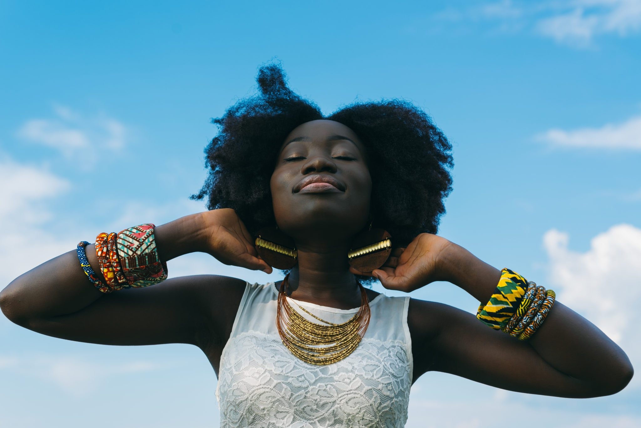 black woman with natural hair looking up to sky with her eyes close while gold bracelets on both arms