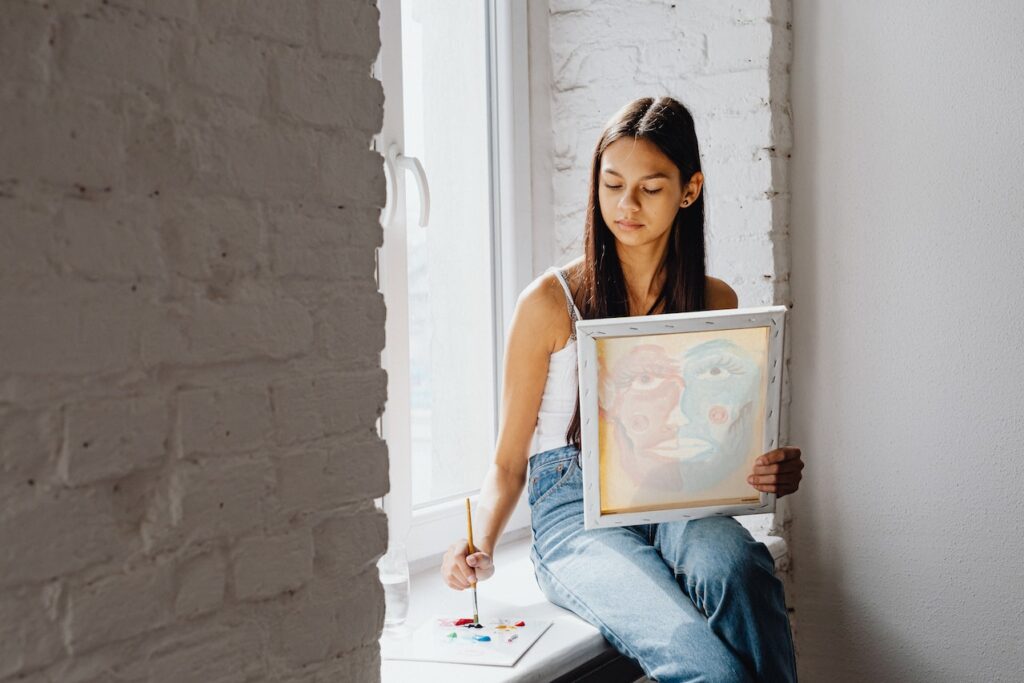 Latine youth painting on a window sill, holding her canvas
