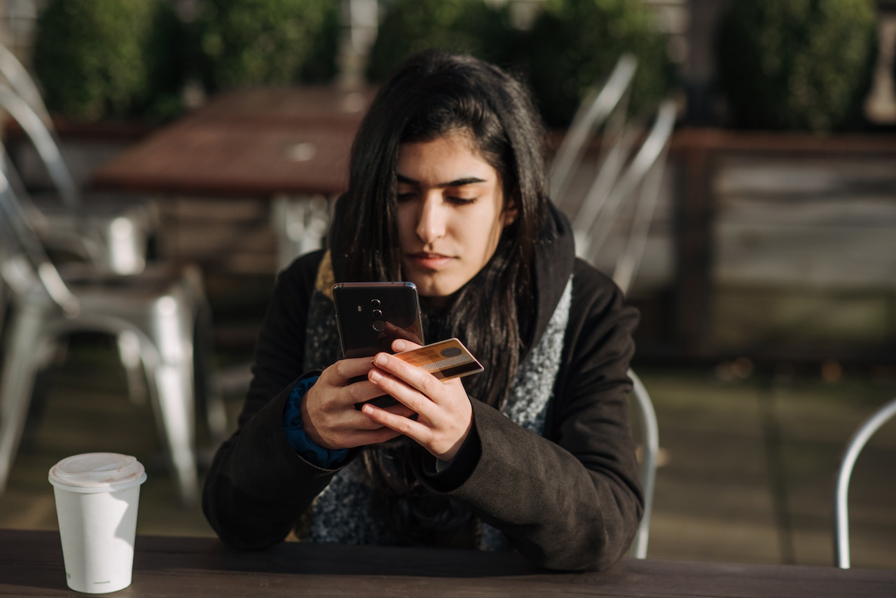 college student sitting at outdoor table looking phone while holding credit card in hand