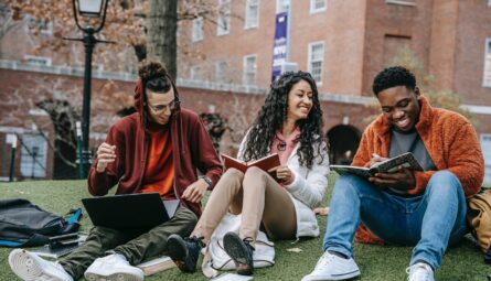 three students sitting on campus lawn talking. reading books and looking at a laptop