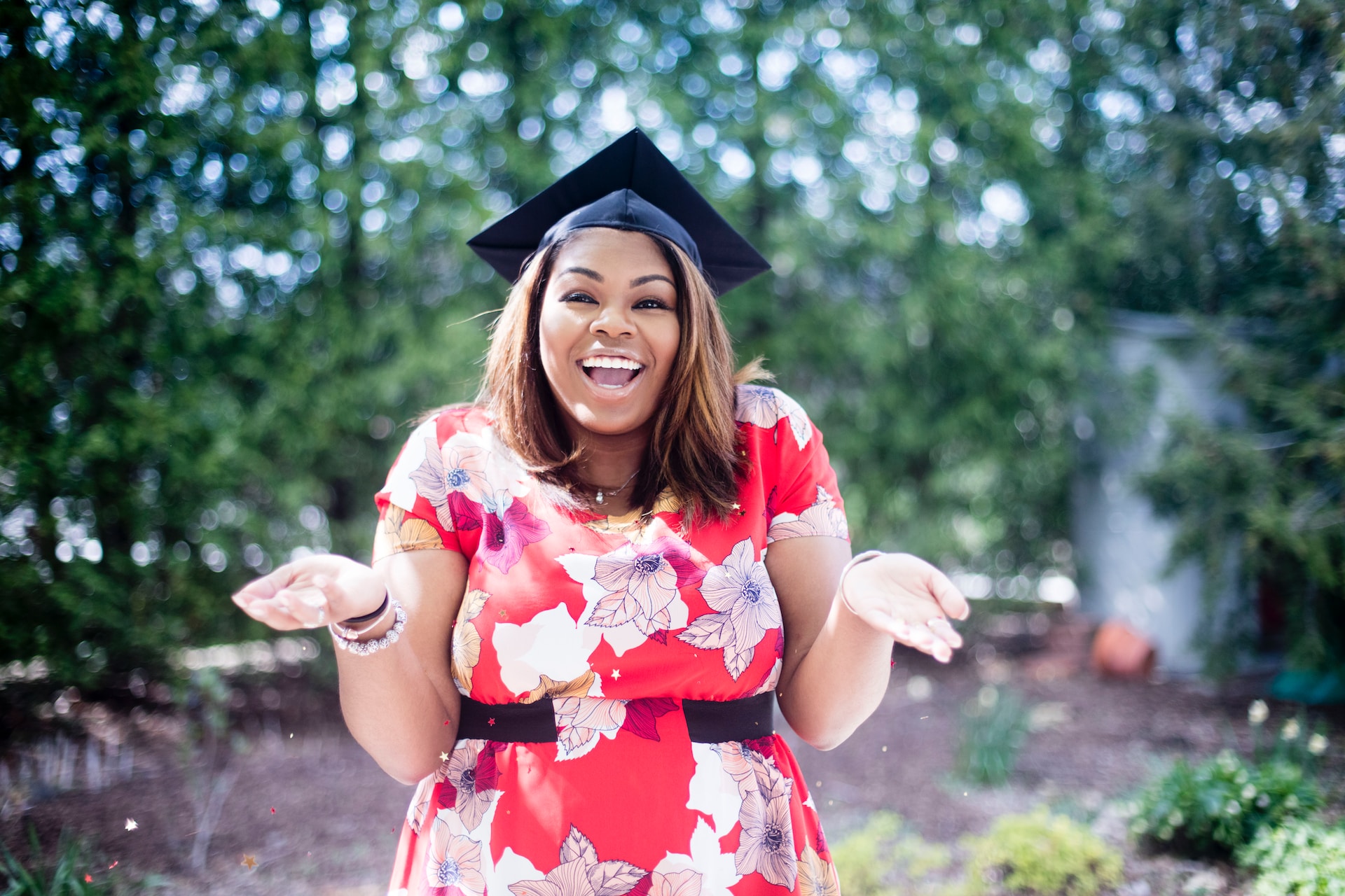 smiling recent college graduate excited about the career skills they have developed. Student is standing outside and wearing floral dress with her arms stretched out and hands facing upward