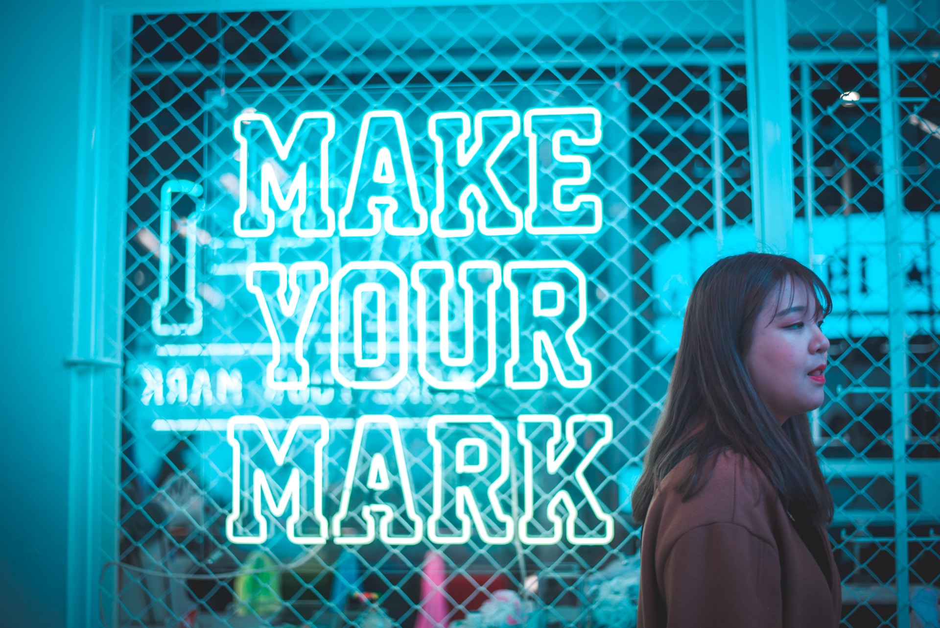 young adult walking passed a neon blue light sign that says make your mark