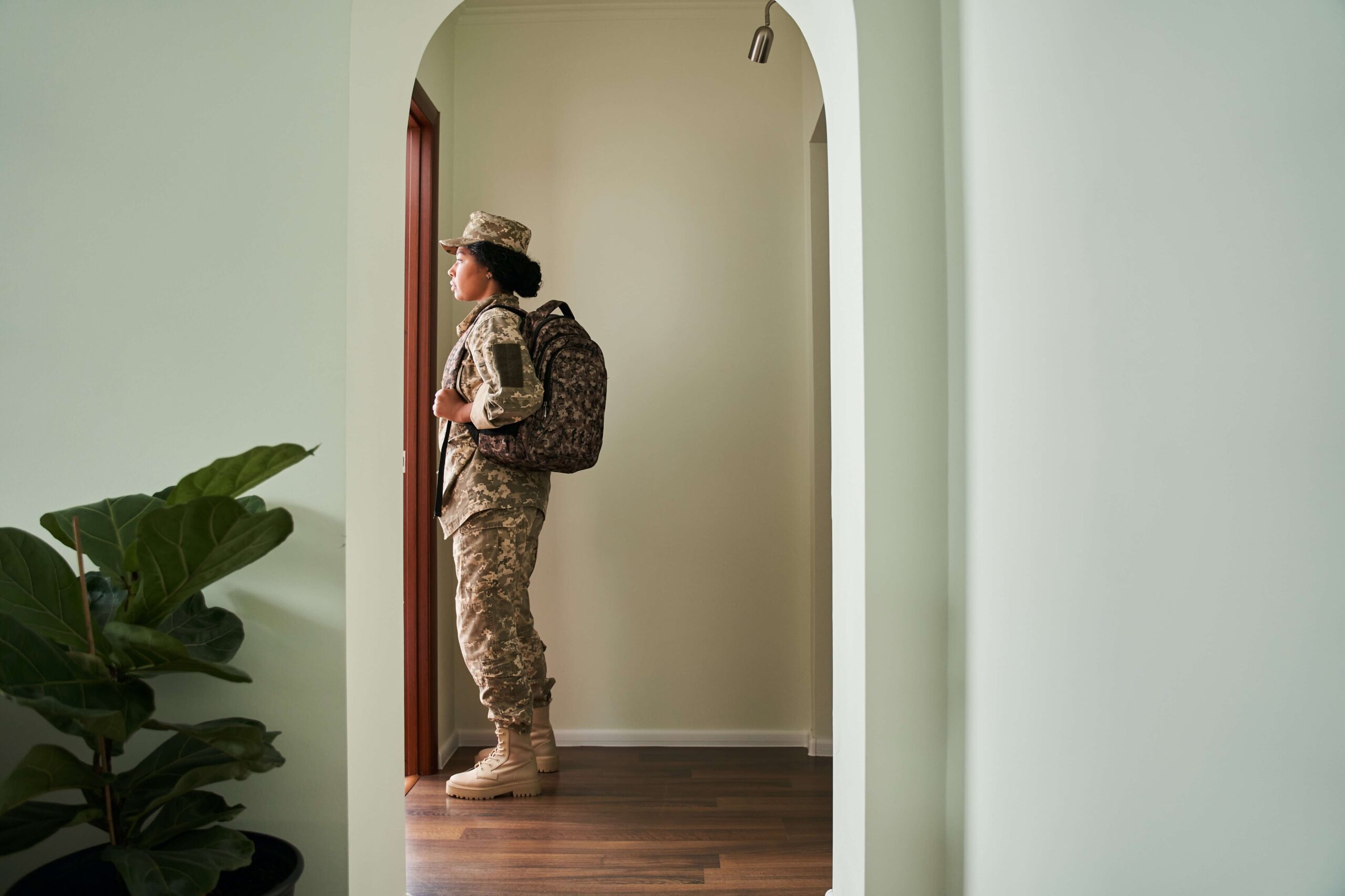 A service member in uniform wears a backpack and stands in front of a door, getting ready to leave the house.