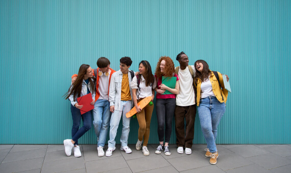 A group of students stands against a blue wall as they smile and look at each other.