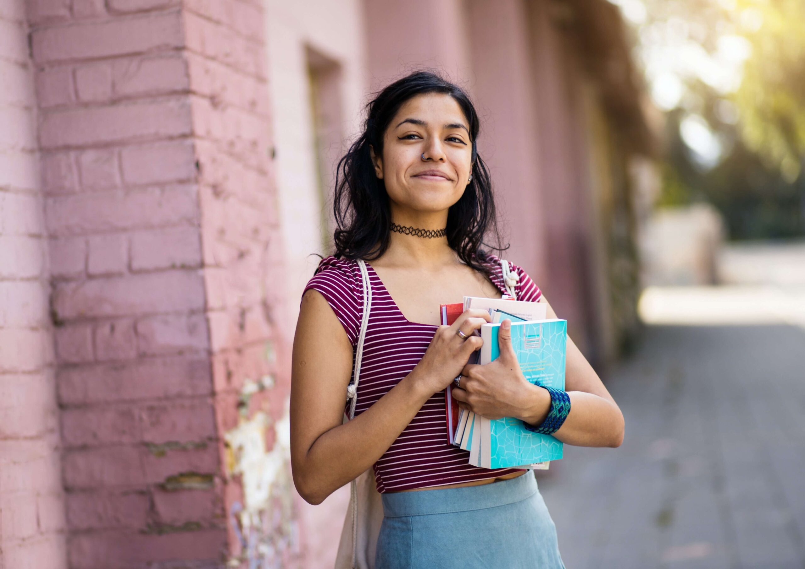 A student smiling outside and holding a stack of books on a sunny day.
