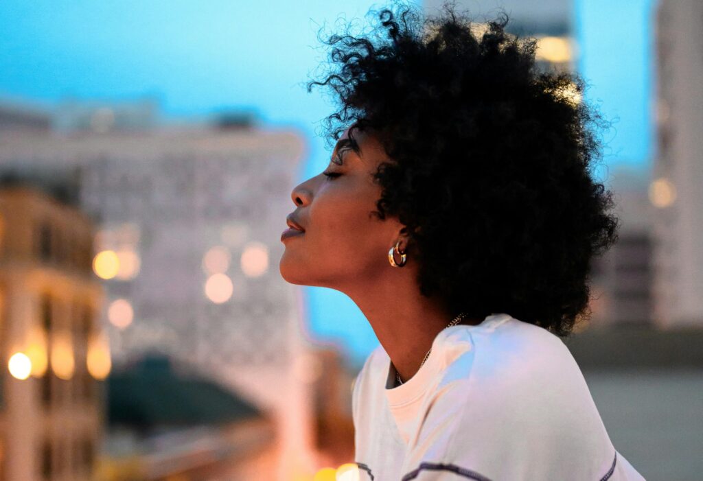A Black woman smiles to herself while closing her eyes as she stands on a balcony on a beautiful night in a city.