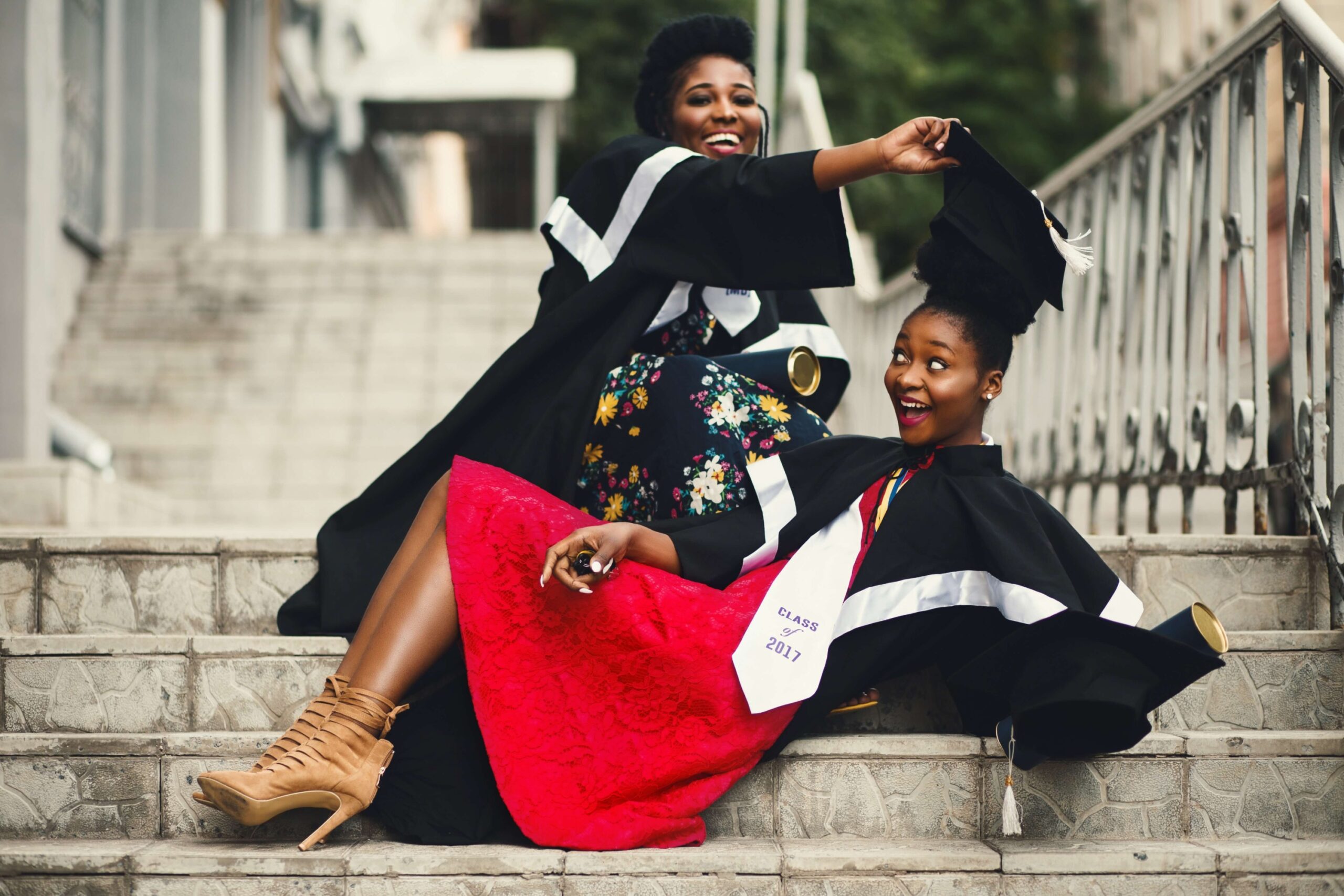 Two Black students wearing graduation gowns smile as one pulls a graduation cap off the other.