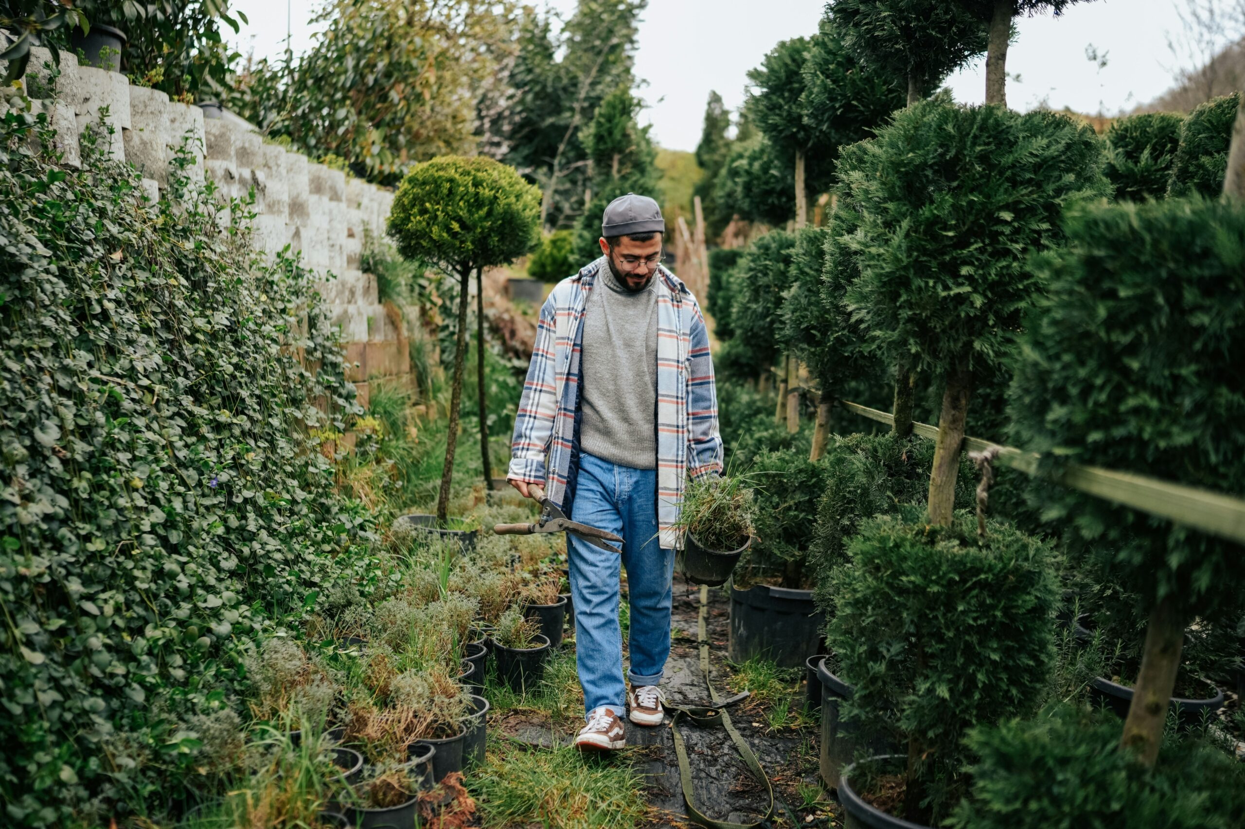 A young person walks through a garden while carrying shears and a plant and looks down with a slight smile on his face.