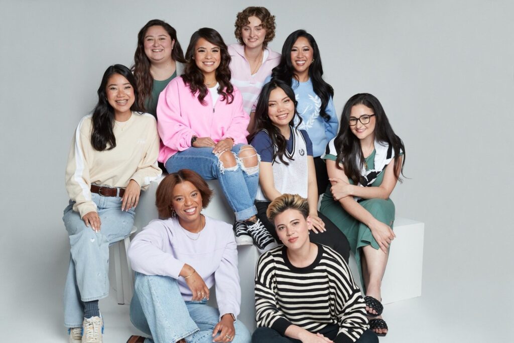 Group of nine diverse young adults posing together in casual, colorful clothing against a light gray studio backdrop. They are smiling and seated or standing on white blocks.