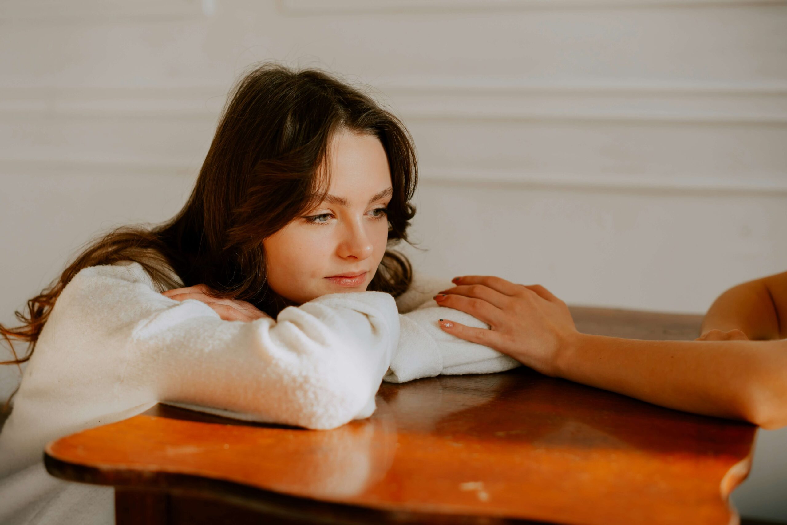 A teen in low spirits rests their head on a table with their arms crossed while a loved one reaches out to offer support.