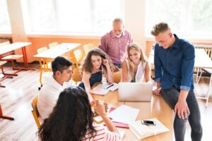 teacher sitting on a desk speaking with a group of students working on a project together