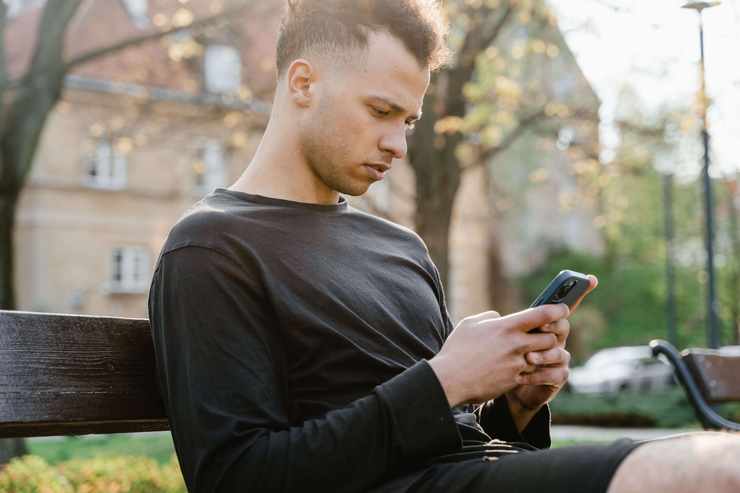 A young man sits on a bench outside and looks at his phone